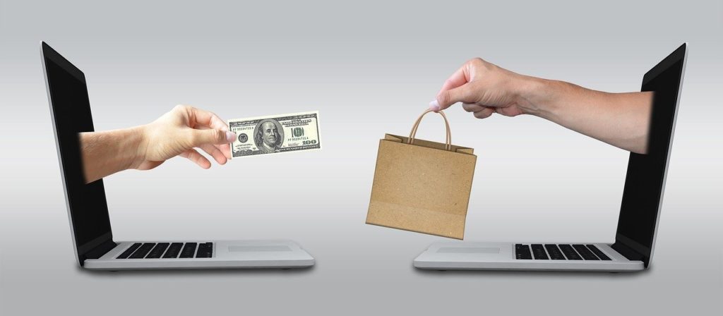 first online business. Two laptops open facing each other. A hand with a dollar bill comes out of one and a hand with a shopping bag from the other. Symbolises ecommerce