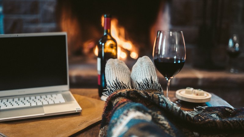 the internet life. A person sits with their laptop in front of a roaring fire with a glass of wine at hand. They are living the internet life  